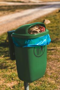 A green trash bin overflowing with garbage in a park during the day, highlighting environmental concerns.