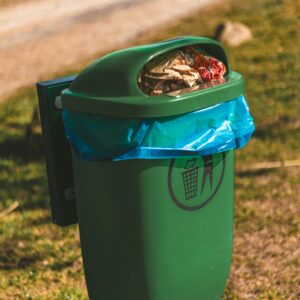 A green trash bin overflowing with garbage in a park during the day, highlighting environmental concerns.