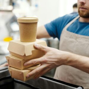 Man wearing apron stacks eco-friendly food containers for takeaway delivery in a kitchen setting.