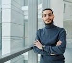 A confident man in a gray turtleneck stands with arms crossed by an office window, exuding professionalism.